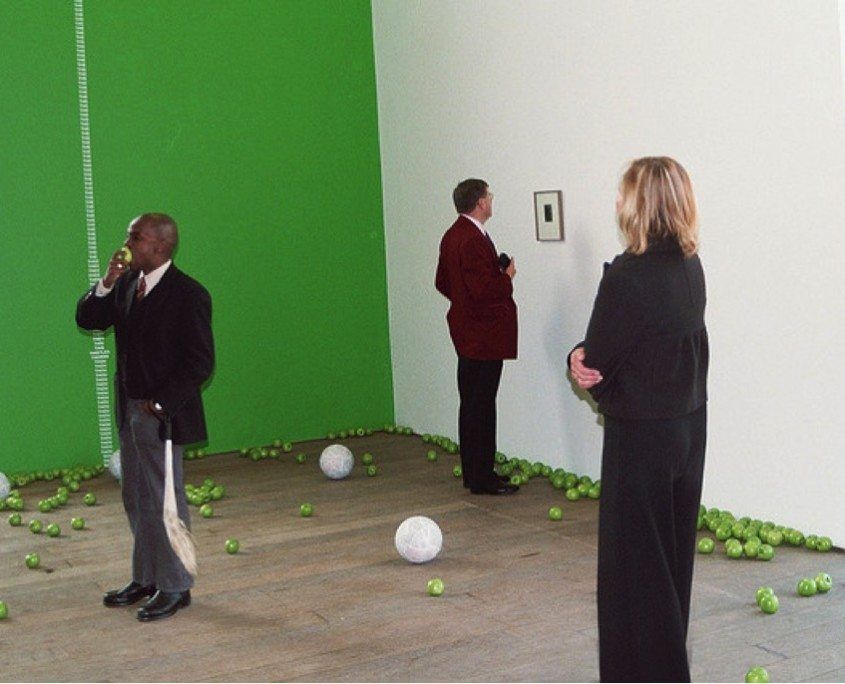 Fig. 2, Samson Kambalu (left) eating an apple and carrying a fly-whisk with patrons viewing (Bookworm) The Fall of Man (2003).