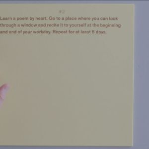 A square, pale yellow sheet of paper rests on a white surface. A hand appears left of the sheet, as though about to turn it over. The light brown text at the top of the sheet reads: “#2 Learn a poem by heart. Go to a place where you can look through a window and recite it to yourself at the beginning and end of your workday. Repeat for at least 5 days.”
