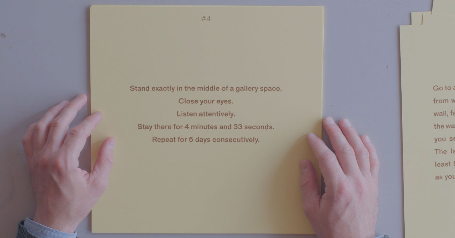 A square, pale yellow sheet of paper rests on a white surface. Two hands are shown on either side of the sheet, palms down. The light brown text on the sheet reads: “#4 Stand exactly in the middle of a gallery space. Close your eyes. Listen attentively. Stay there for 4 minutes and 33 seconds. Repeat for 5 days consecutively.” Each sentence is given its own line.