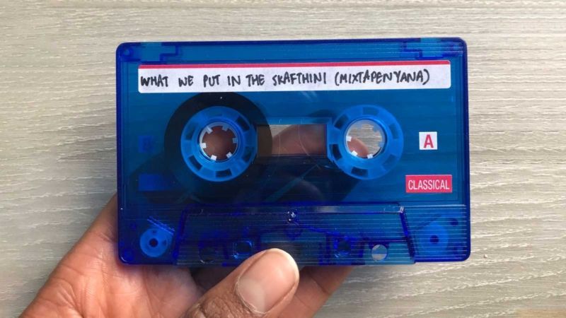The artists hand appears holding a blue, slightly transparent audio cassette tape. On the top of the tape on a white label, the work title, “WHAT WE PUT IN THE SKAFTHINI (MIXAPENAYANA)” appears written in all-caps in black marker.